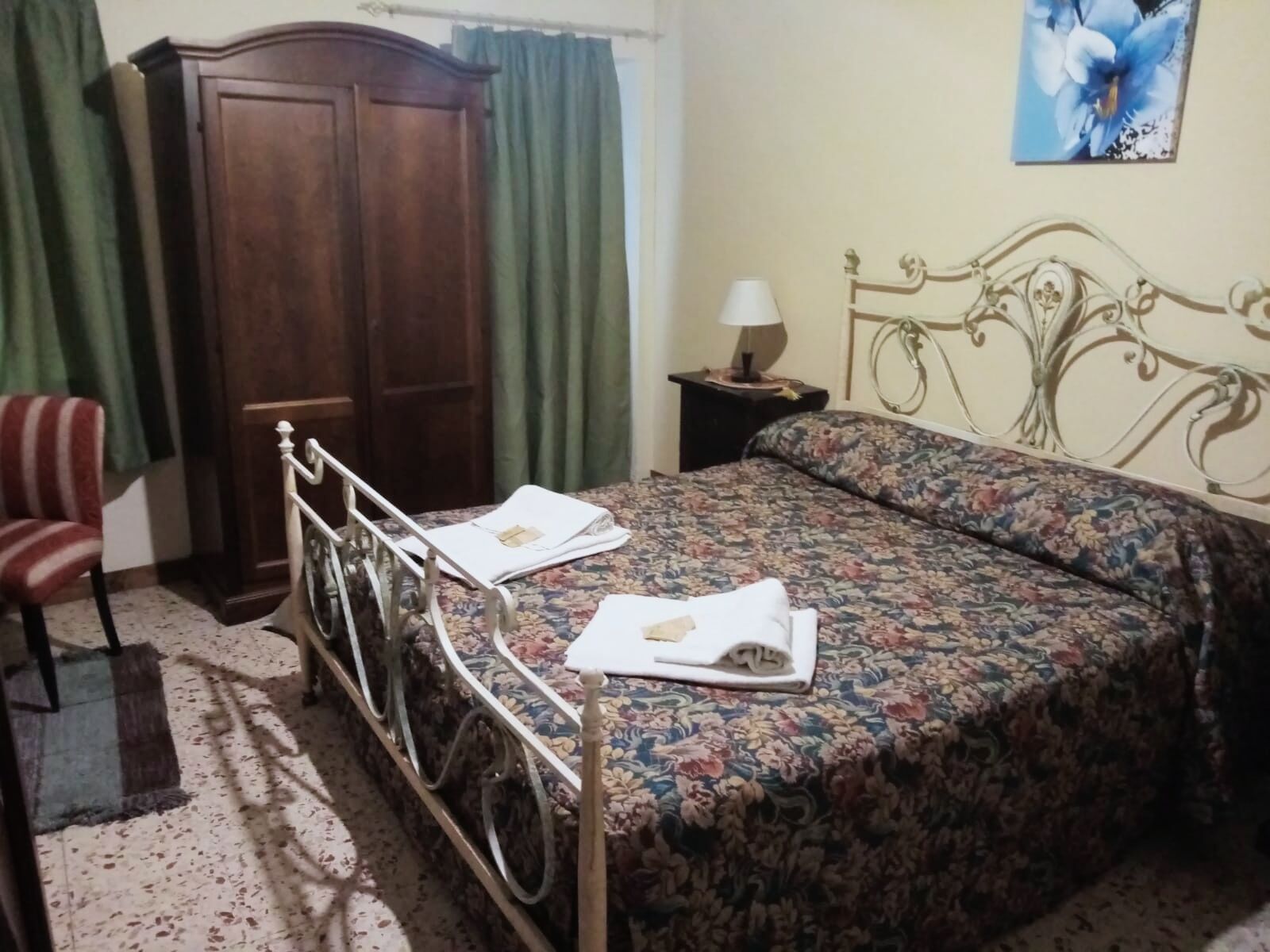 Ein Schlafzimmer mit Rollstuhl, einem Bett mit Blumenmuster auf der Bettwäsche, einem Nachttisch mit Lampe, einem Kleiderschrank und einem Sessel.