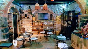 Un interior acogedor y rústico con paredes de ladrillo, un arco de piedra y un televisor sobre la chimenea. Varias mesas y sillas están dispuestas para comer, y se exhiben diversos objetos decorativos y plantas en todo el espacio.