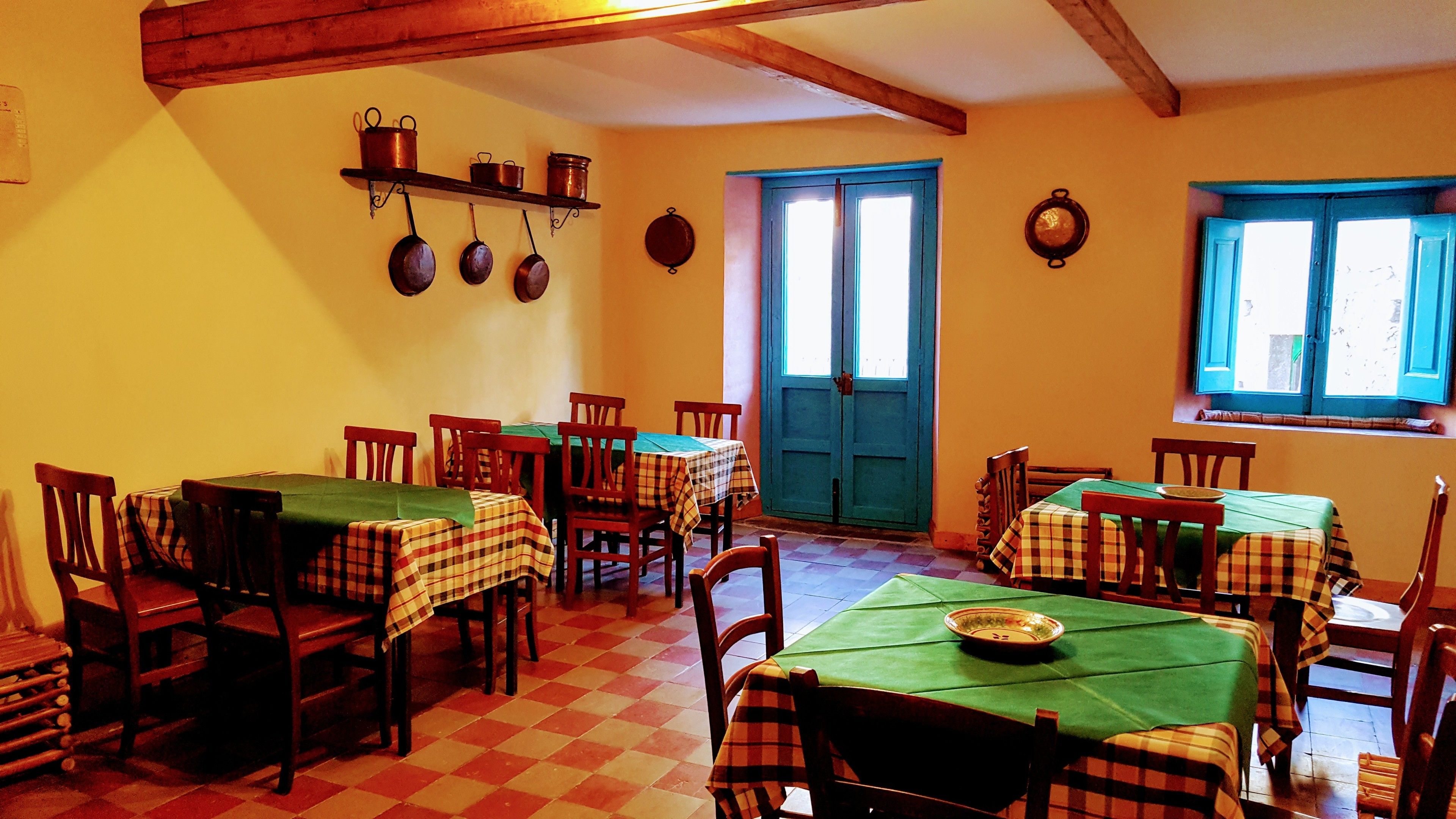 A dining area with checkered floor tiles, wooden tables with plaid tablecloths, and chairs. There are hanging pots on a shelf above the tables.
