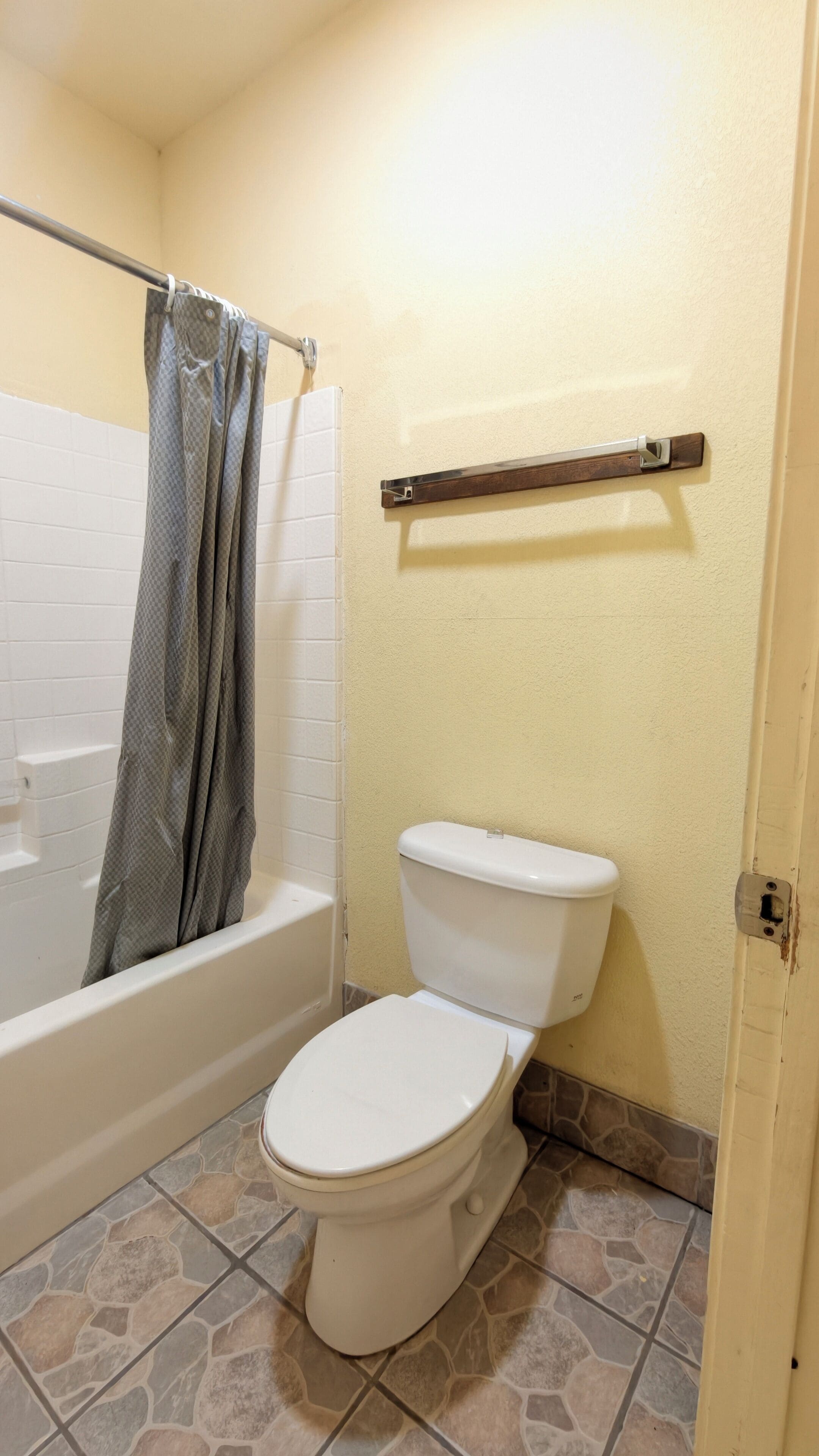 A bathroom with a white toilet, a shower with a grey curtain, a wooden shelf, and a tiled floor.