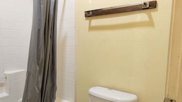 A bathroom with a white toilet, a shower with a grey curtain, a wooden shelf, and a tiled floor.
