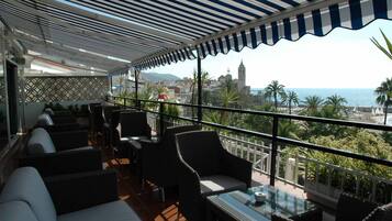 Une terrasse dotée de meubles en osier, d’une table en verre et offrant une vue sur la mer et le paysage urbain.