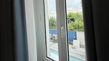 Ett rum med ett stort fönster, gardiner och utsikt över en swimmingpool och byggnader utanför.