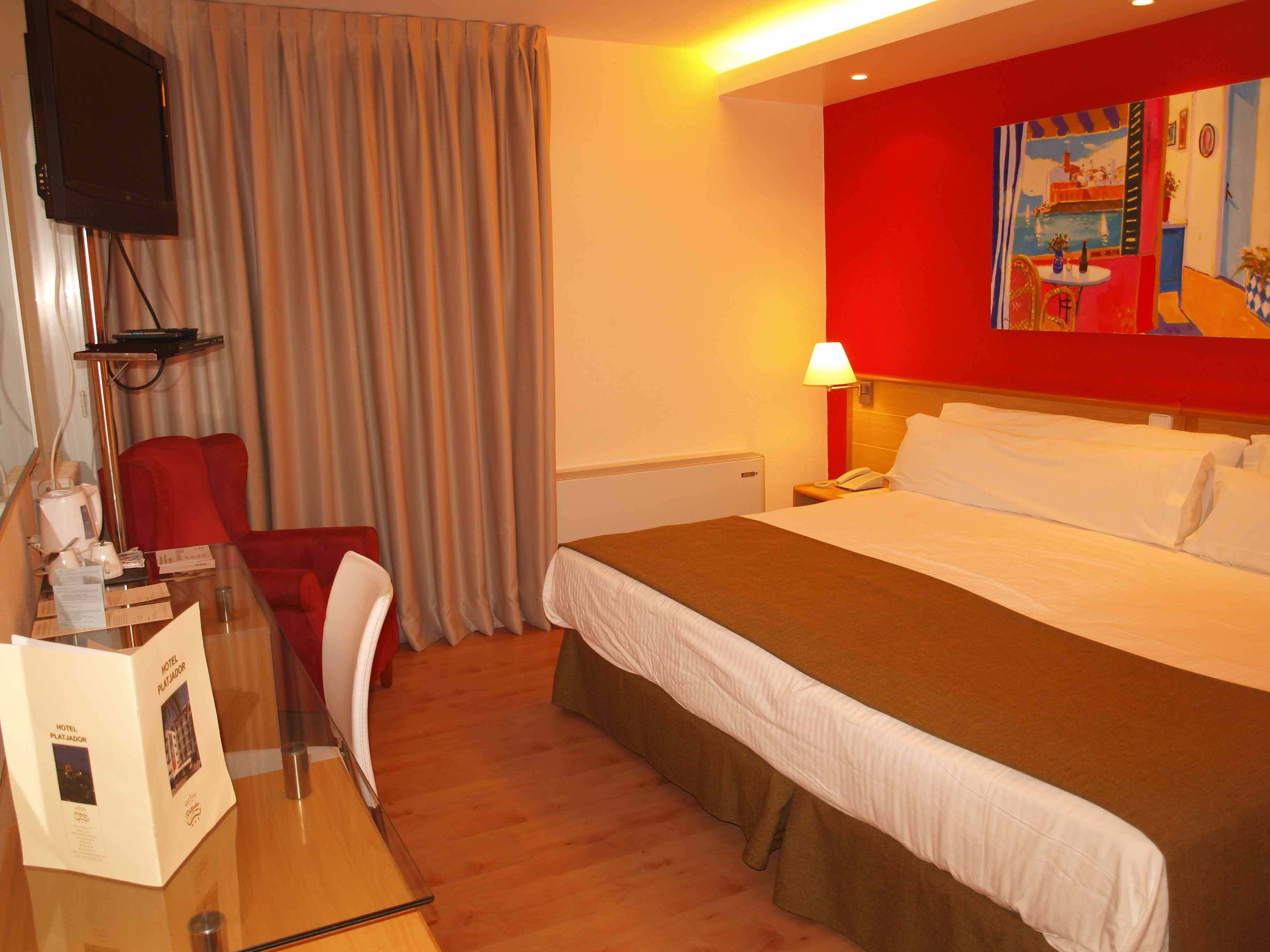 Una habitación de hotel con una cama, un escritorio con silla, un televisor y un cuadro en la pared.