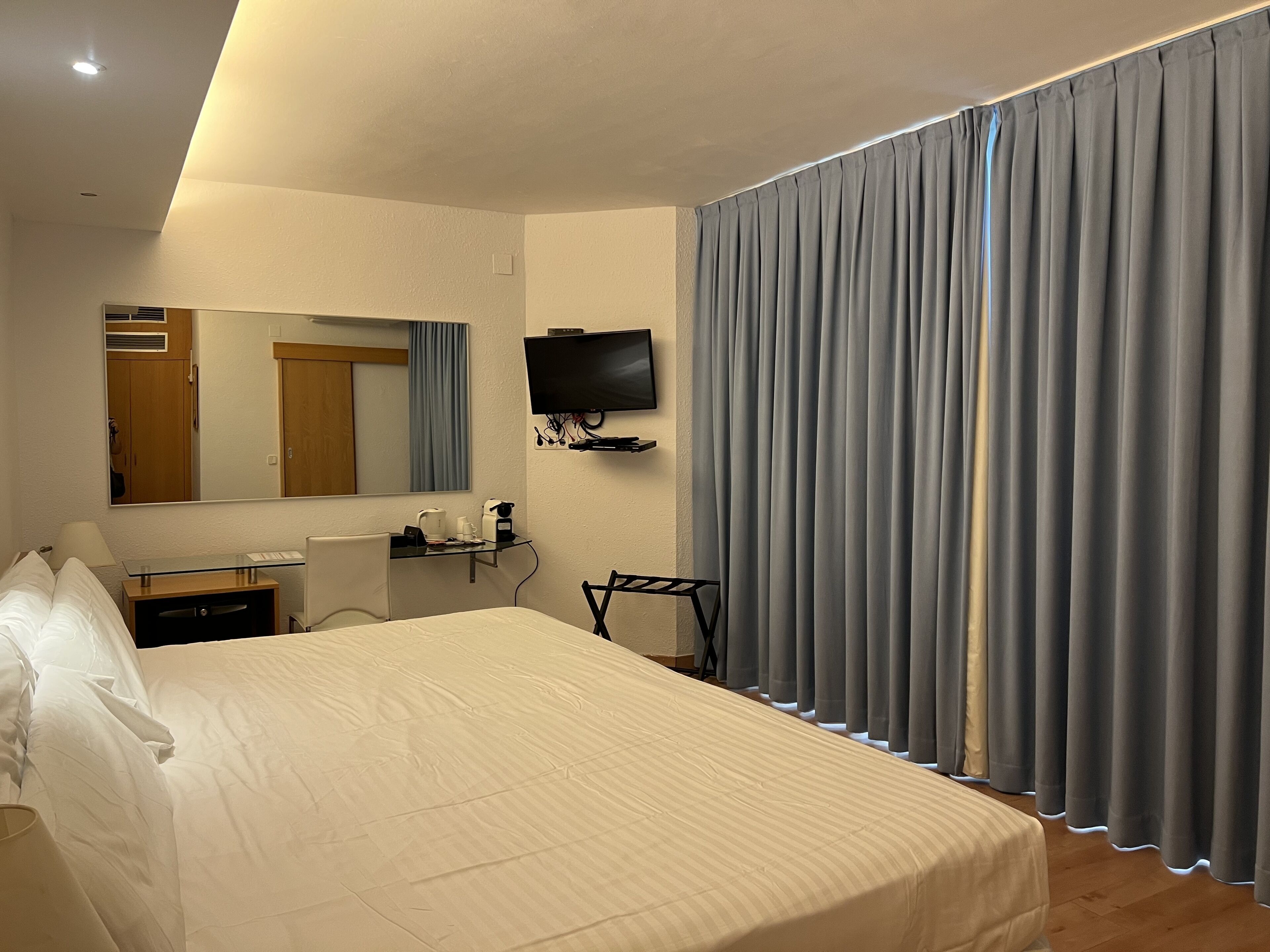 A hotel room with a bed, a desk with a chair, a television, a mirror, and a window with curtains.