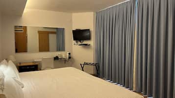 A hotel room with a bed, a desk with a chair, a television, a mirror, and a window with curtains.
