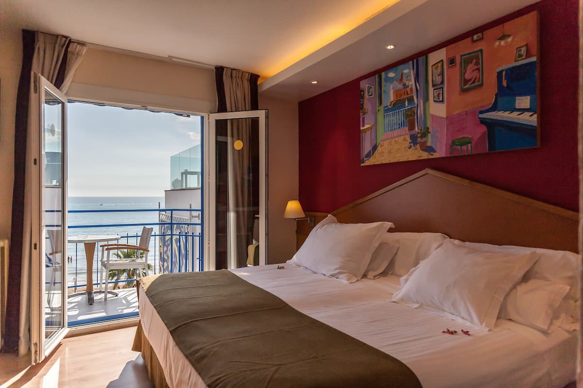 Ein Hotelzimmer mit Bett, Meerblick, einem Gemälde an der Wand und einem Balkon mit Tisch und Stühlen.