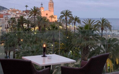 A terrace with wicker furniture and a candle, overlooking a coastal cityscape with palm trees and a prominent building.