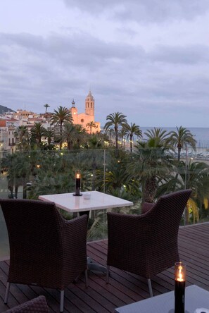 A terrace with wicker furniture and a candle, overlooking a coastal cityscape with palm trees and a prominent building. - Platjador Hotel (Sitges)