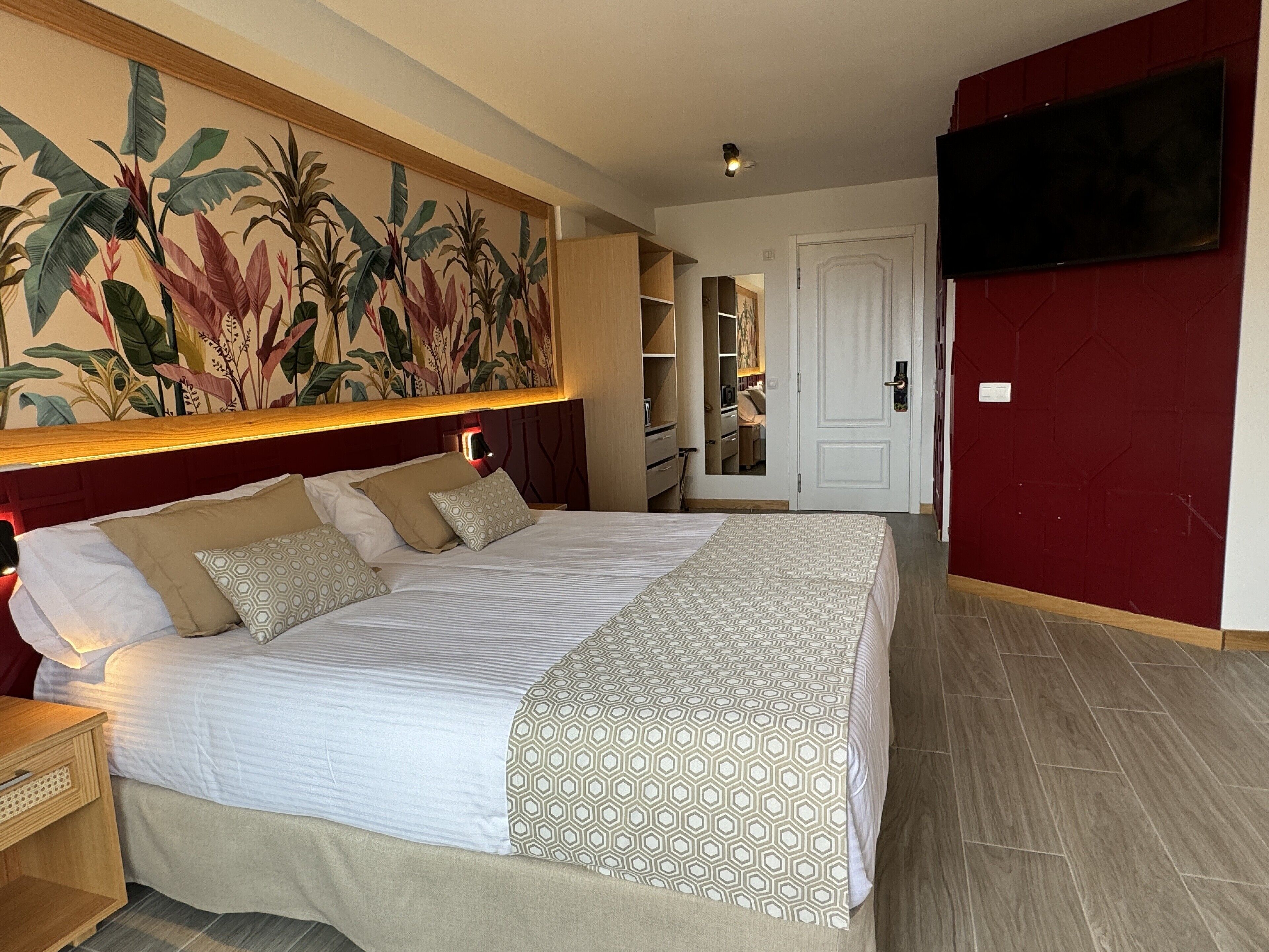 Un lit bien fait, avec une literie à motifs, une grande fresque murale, un téléviseur fixé au mur et un mur d’accent de couleur rouge.