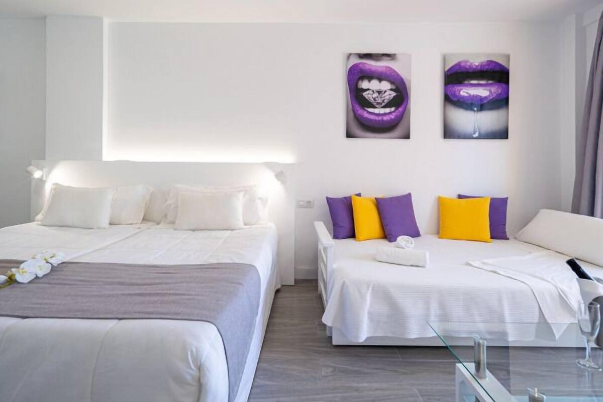 Habitación de hotel con dos camas, ropa de cama blanca, almohadas moradas y amarillas, y una obra de arte en la pared.