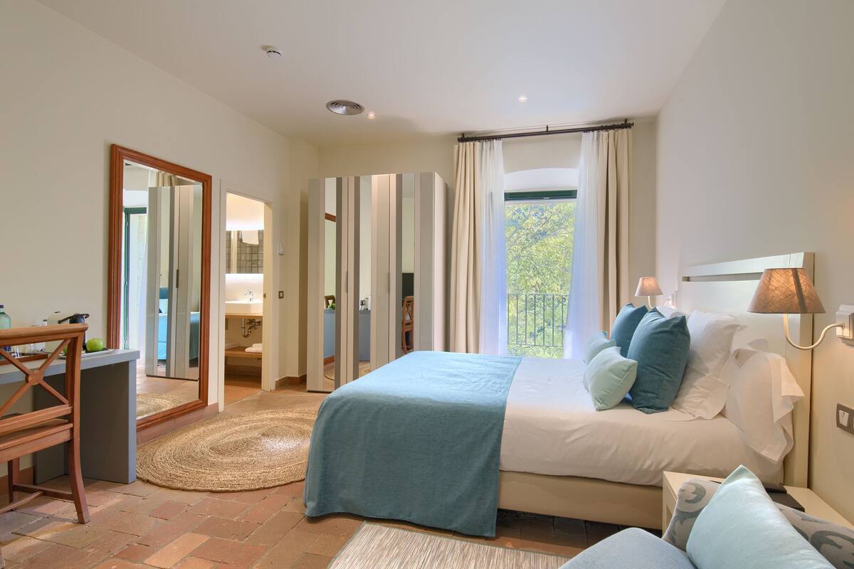 A bedroom with a bed, bedside table, lamp, and a view of the outdoors.