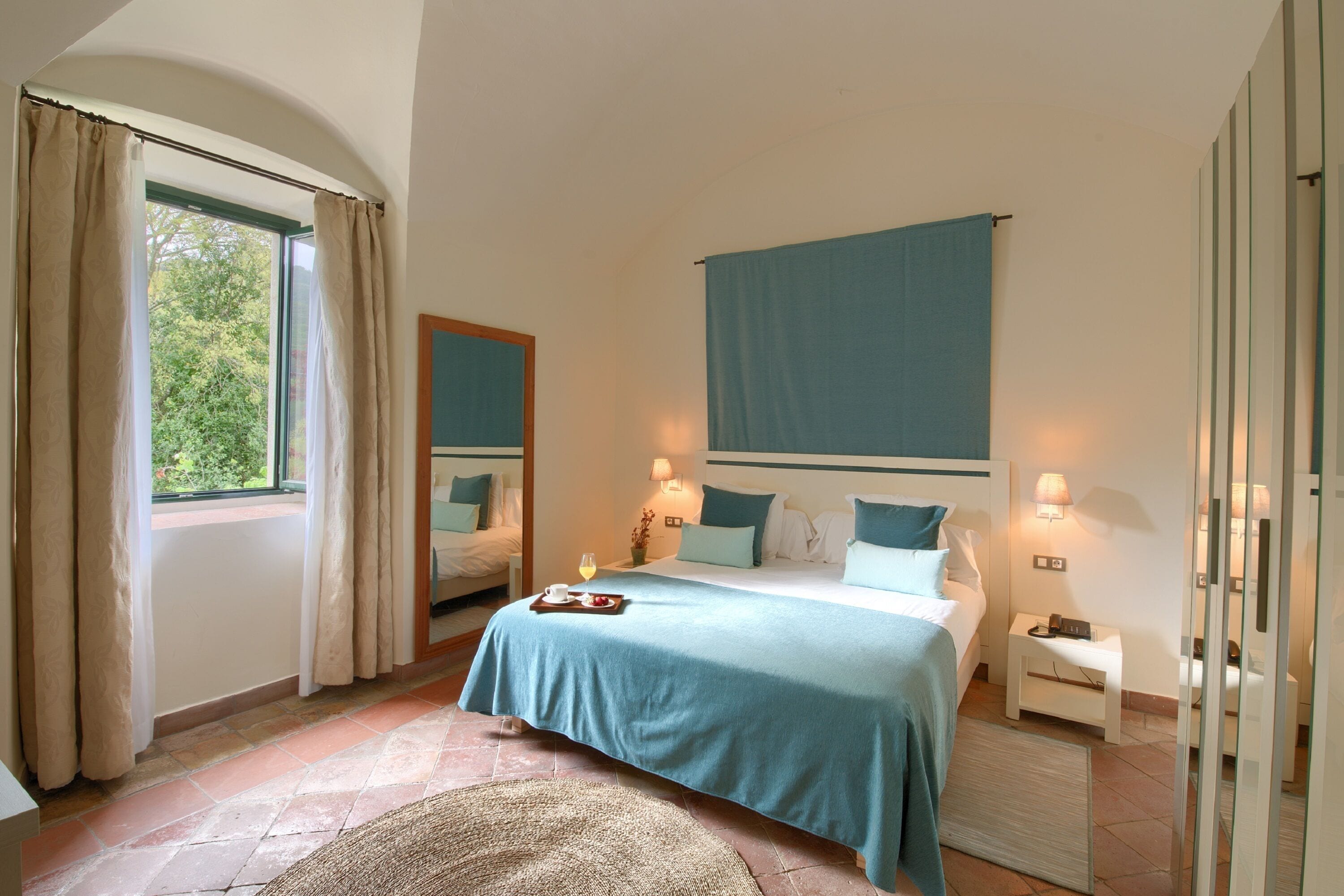 Un dormitorio con cama, mesitas de noche, un tocador, un espejo y una ventana con cortinas.