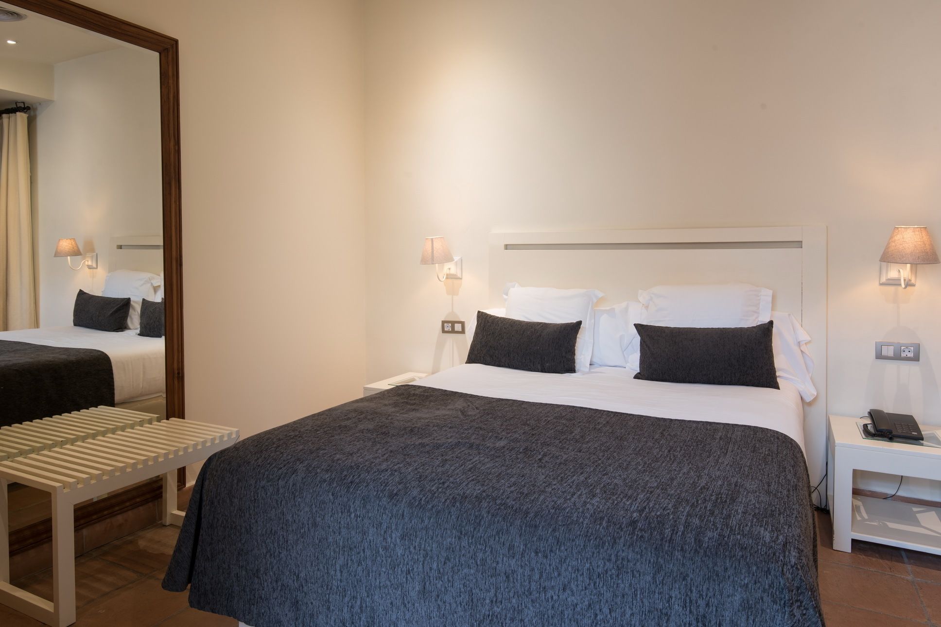 Habitación de hotel con una cama grande, dos mesitas de noche, un espejo y un pequeño rincón de descanso.