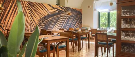 A dining area with wooden tables and chairs, a large wall mural, and a glassware cabinet.