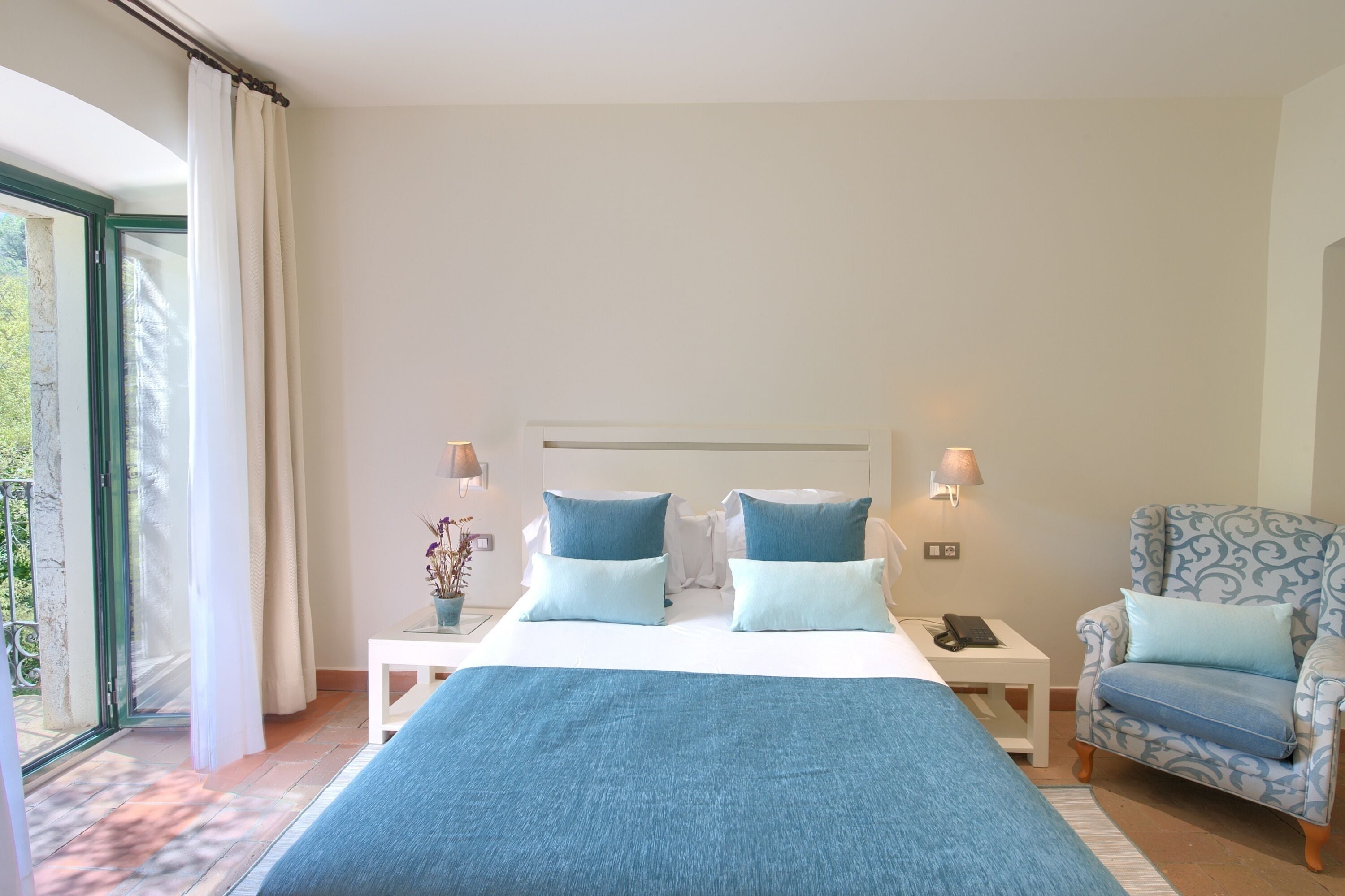 Une chambre à coucher comprenant un lit, deux tables de chevet, une chaise, une lampe et une vue sur la verdure par une fenêtre.