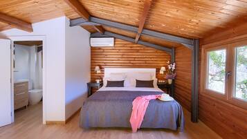 A bedroom with a wooden ceiling, a bed with a pink blanket, a nightstand with a lamp, and a window with a view of greenery.