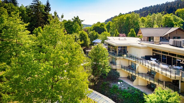 Een hotel met meerdere verdiepingen, met buiten zitplekken, omringd door weelderige begroeiing en bomen.
