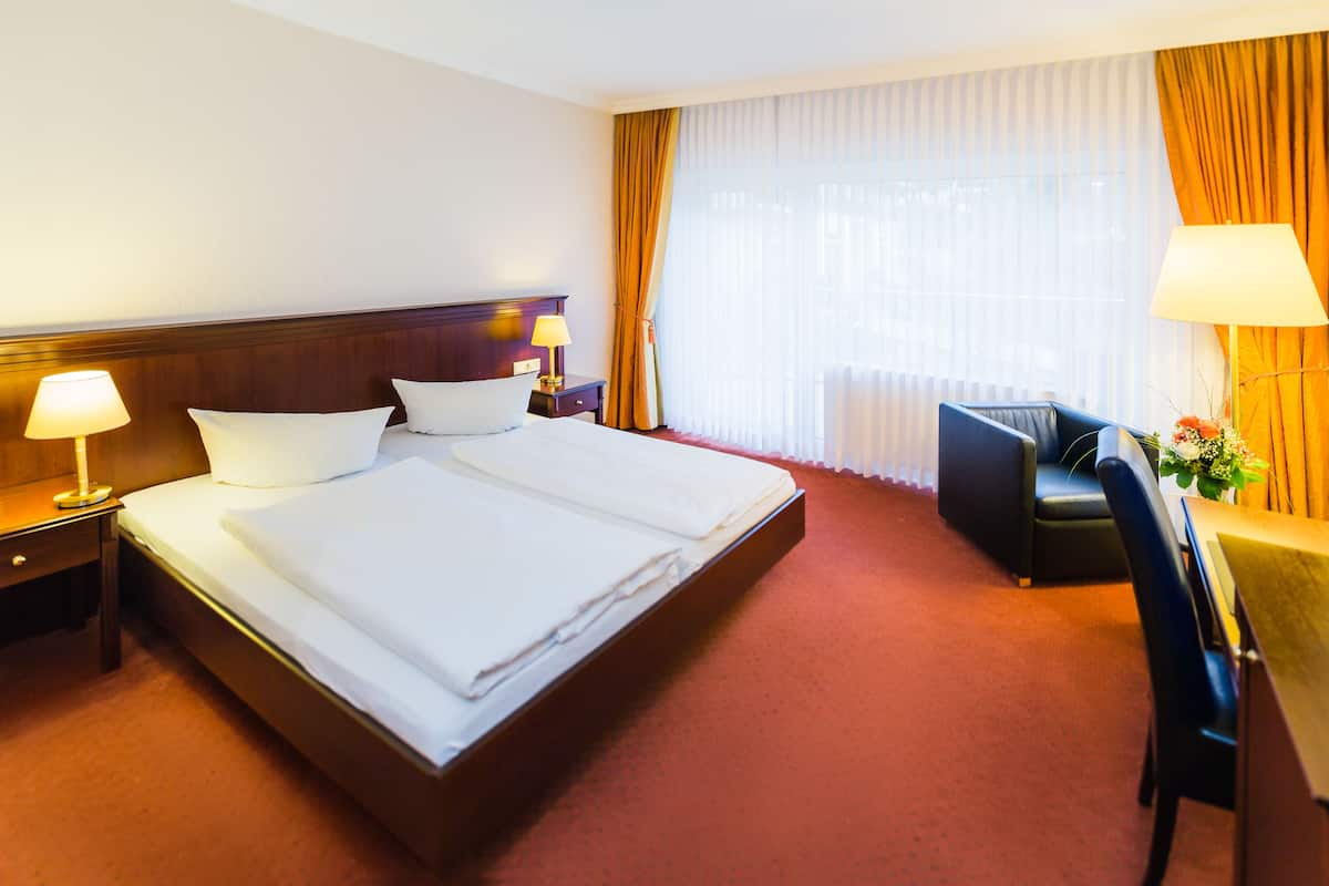 A hotel room with a double bed, two bedside tables with lamps, a desk with a chair, and a large window with curtains.