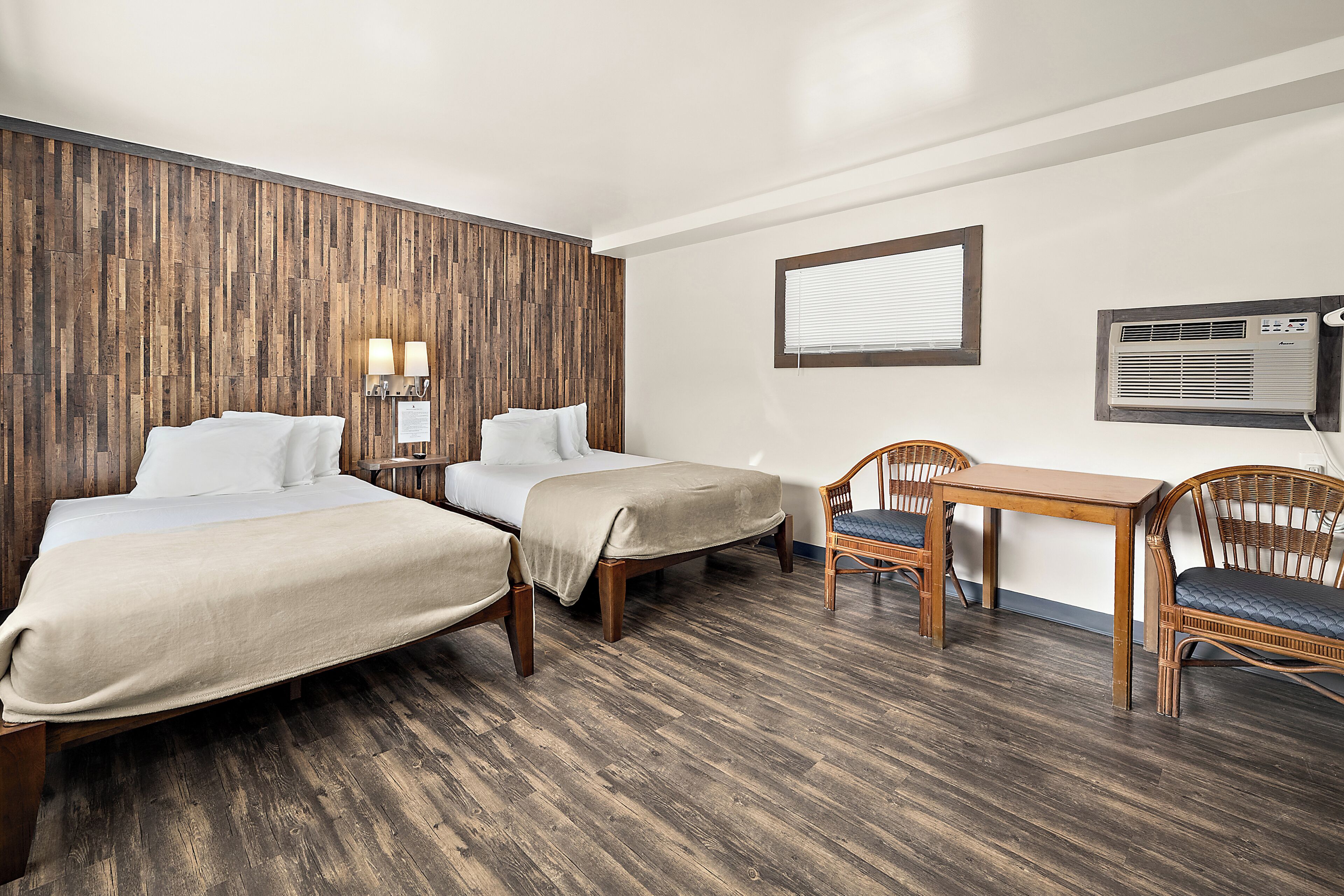 Quarto de hotel com duas camas, uma mesa pequena, uma cadeira, ar condicionado na parede e uma janela com persianas.