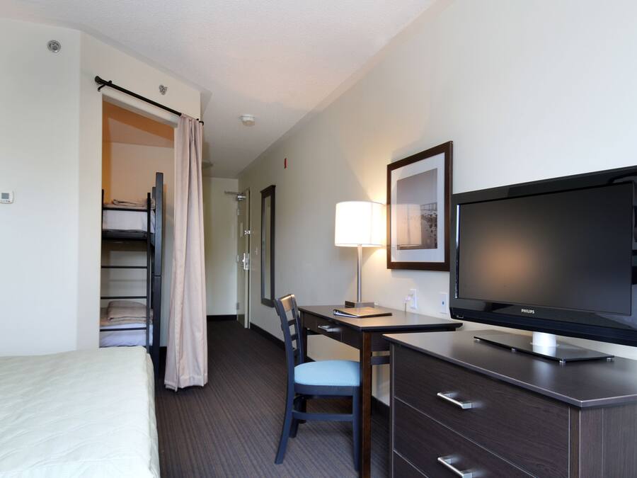 A hotel room with a bed, a desk with a chair, a television, a lamp, and a wardrobe.