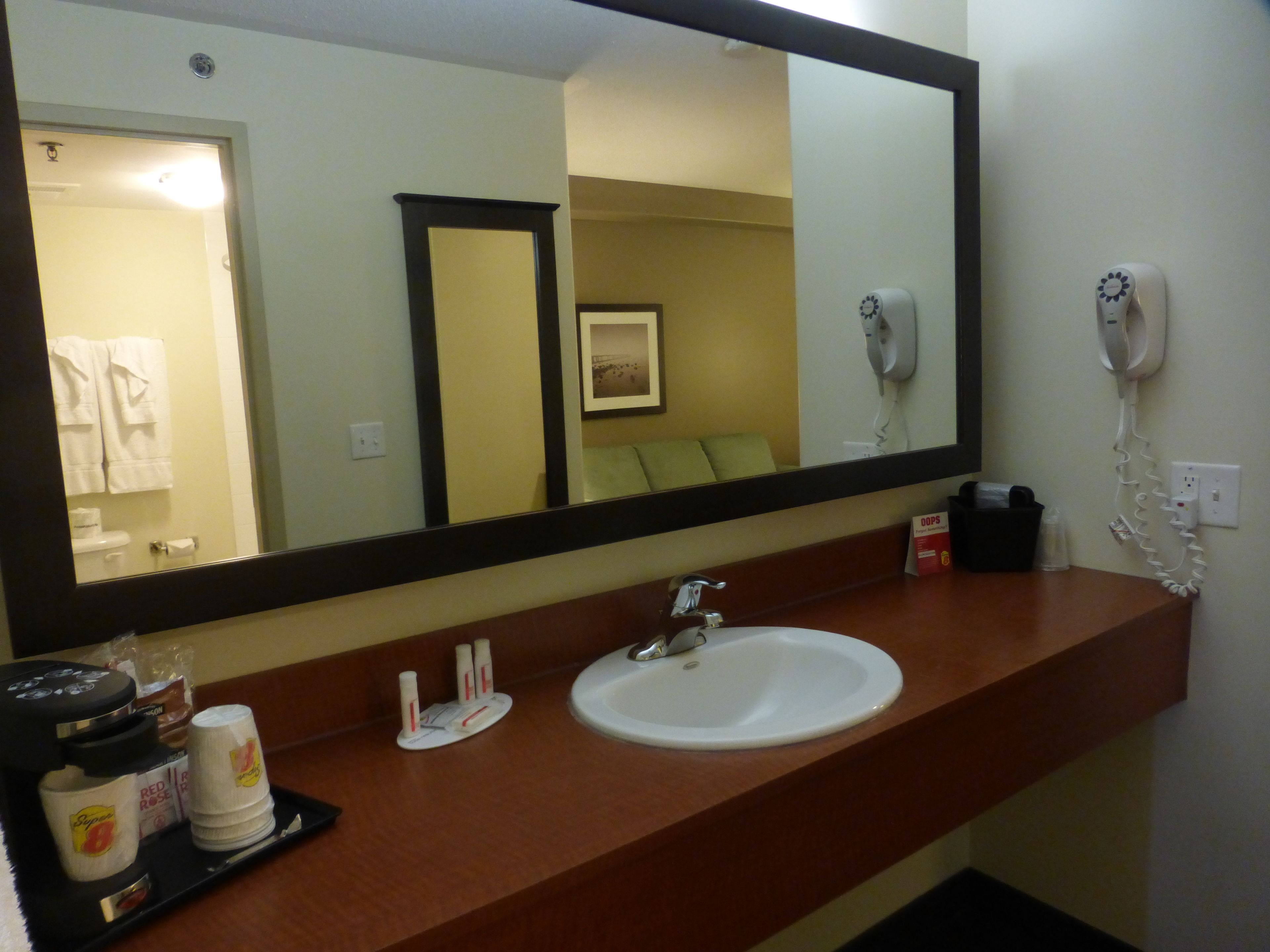 A hotel bathroom with a large mirror, a sink, a hairdryer, and a tray with toiletries.