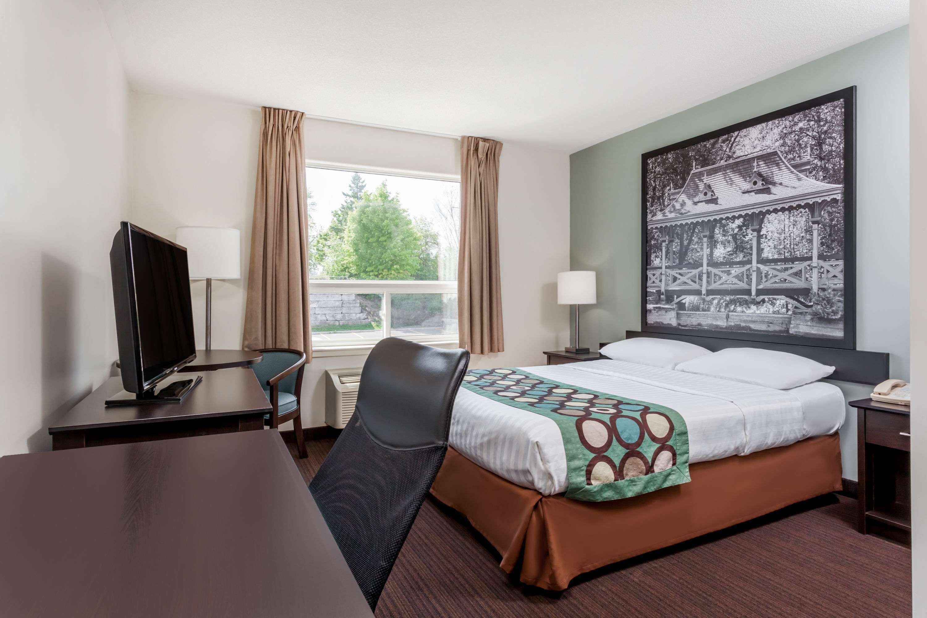 Habitación de hotel con cama, escritorio con computadora, silla, ventana con cortinas, mesita de noche con lámpara y un cuadro en la pared.