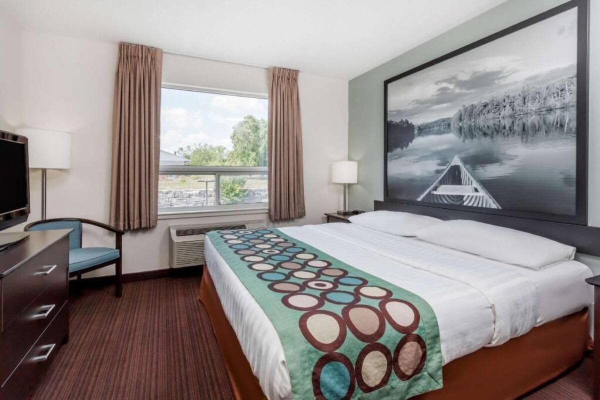 Habitación de hotel con cama, escritorio, silla, televisor, ventana con cortinas y un cuadro grande en la pared.