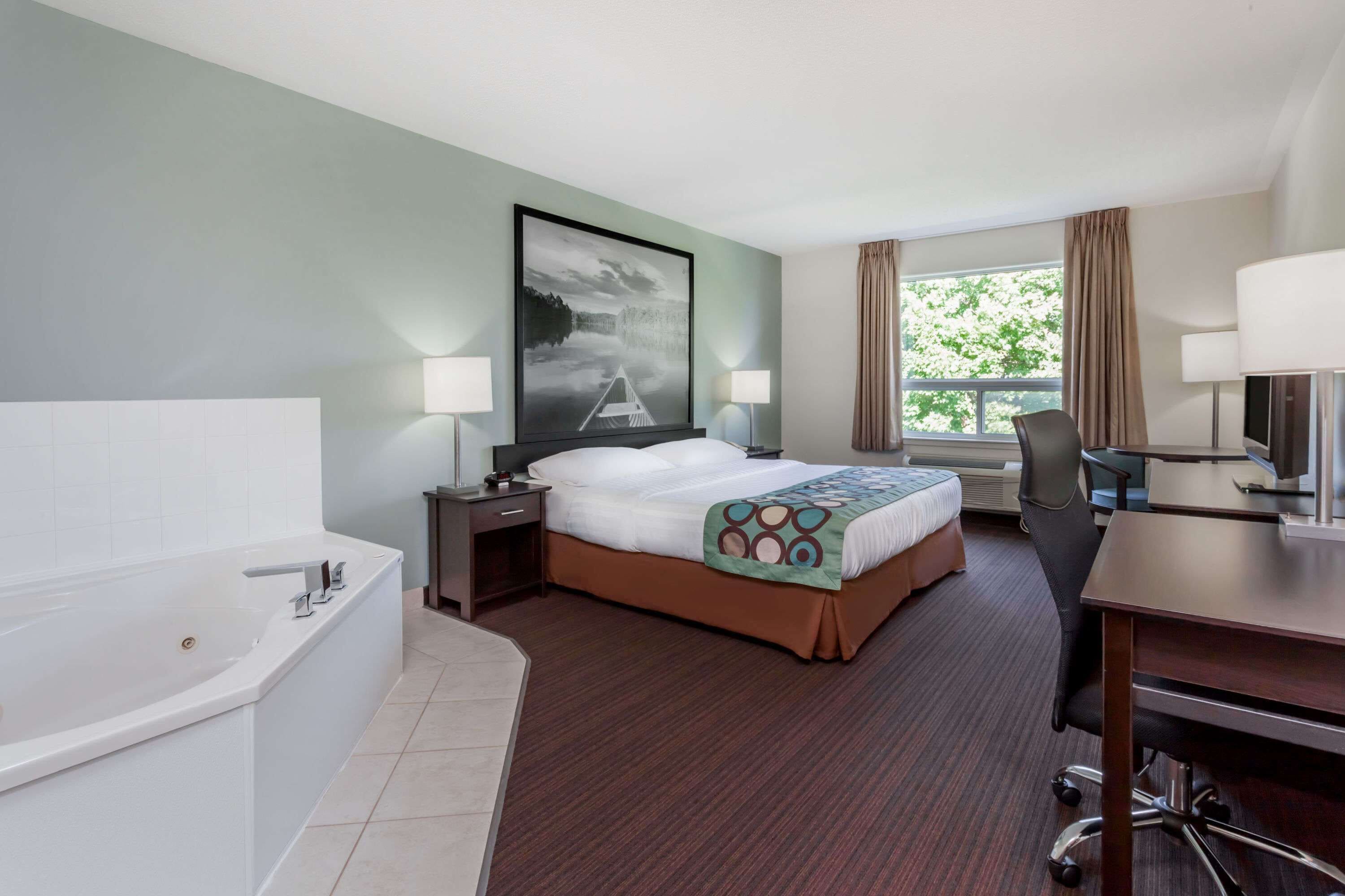 A hotel room with a large bed, a separate desk with a chair, a desk lamp, a large window with curtains, a painting on the wall, and a bathtub.