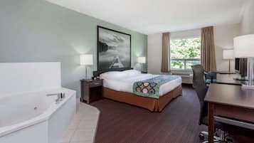 A hotel room with a large bed, a separate desk with a chair, a desk lamp, a large window with curtains, a painting on the wall, and a bathtub.