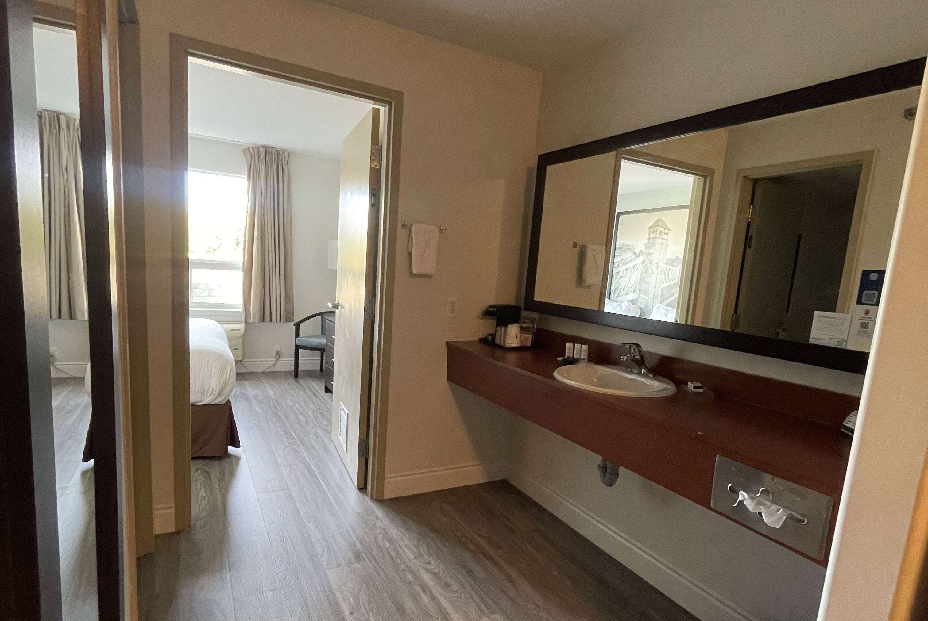 A hotel bathroom with a wooden vanity, a large mirror, and a view of a bedroom with a bed and curtains.