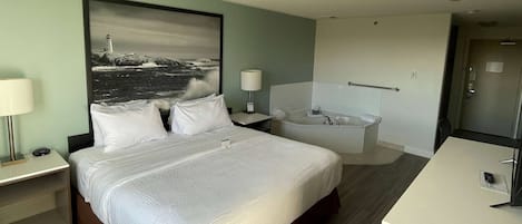 Habitación de hotel con cama, mesita de noche, escritorio, baño con bañera y un cuadro grande de un faro en la pared.