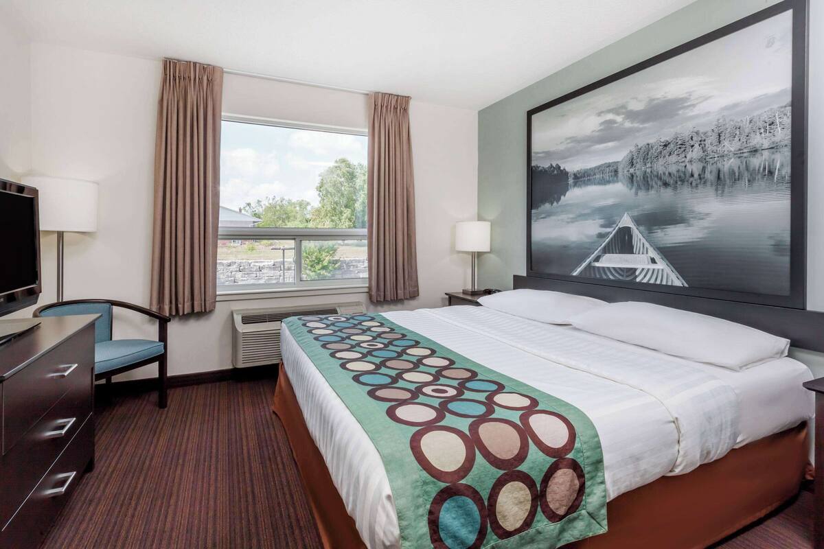 Habitación de hotel con una cama grande, un escritorio, una silla, un televisor, una ventana con cortinas y un cuadro de un lago.