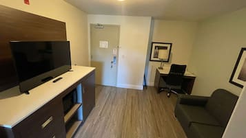 A compact hotel room with a flat-screen TV, a desk, a chair, and a couch.