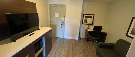 A compact hotel room with a flat-screen TV, a desk, a chair, and a couch.