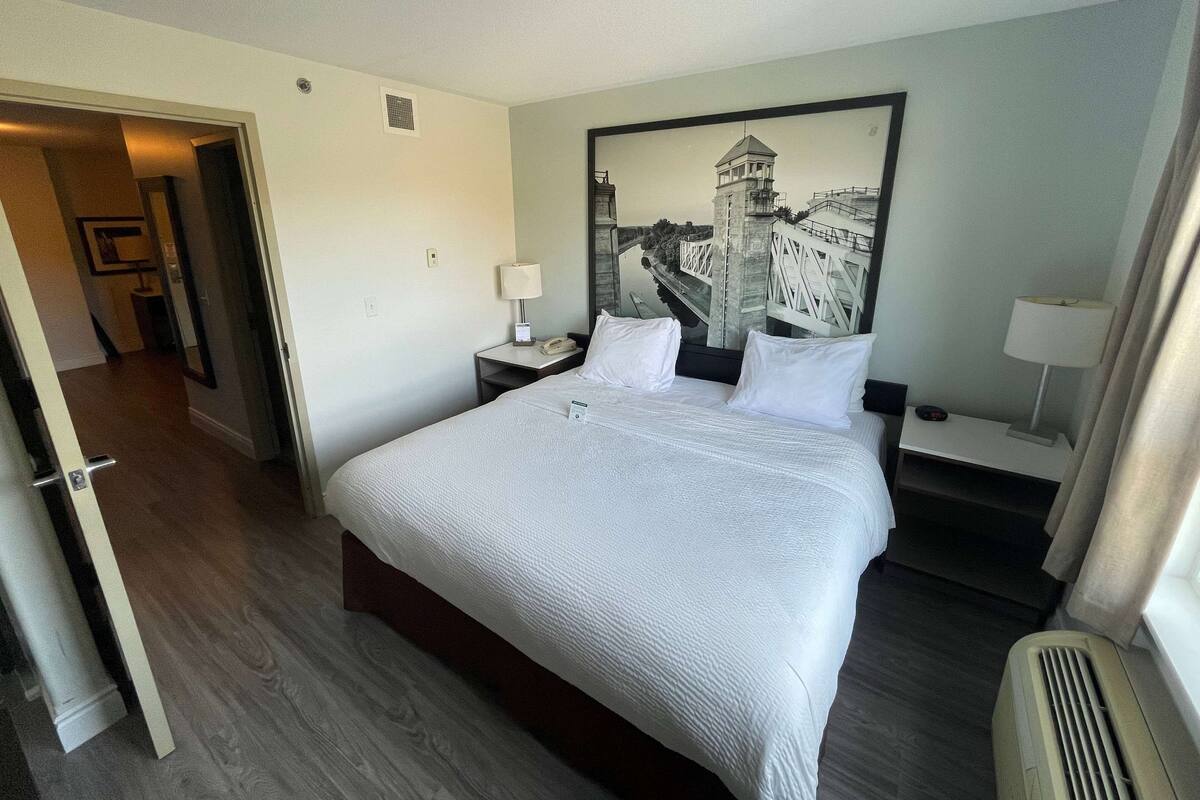 Habitación de hotel con una cama grande, una mesita de noche con lámpara, un cuadro en la pared y una puerta que da a otra habitación.