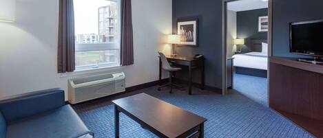 Una habitación de hotel con un sofá azul, una mesa de madera, un escritorio con una silla, un televisor y un amplio ventanal con cortinas.