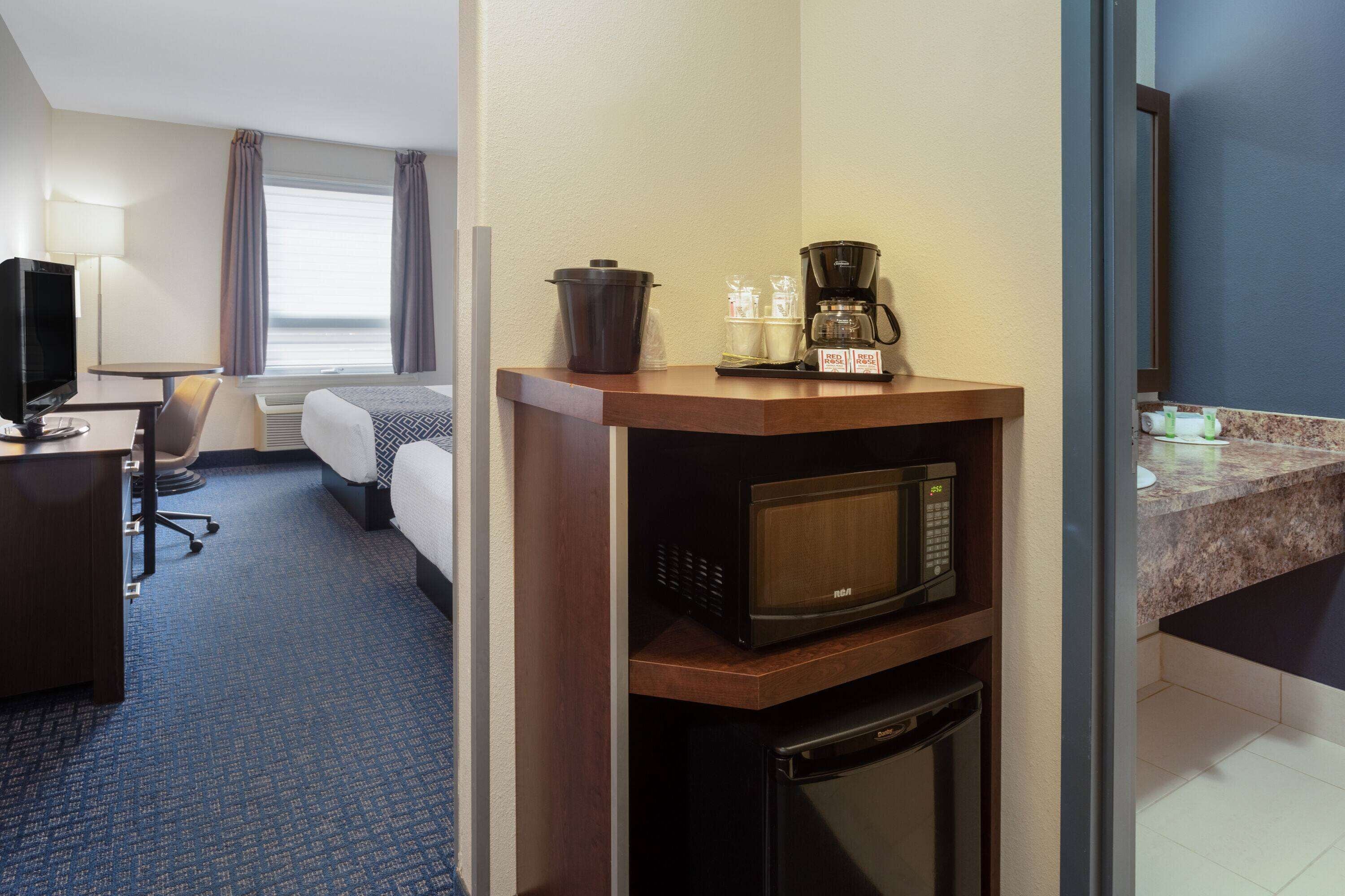 A hotel room with a kitchenette, a bed, a desk with a chair, a television, and a window with curtains.