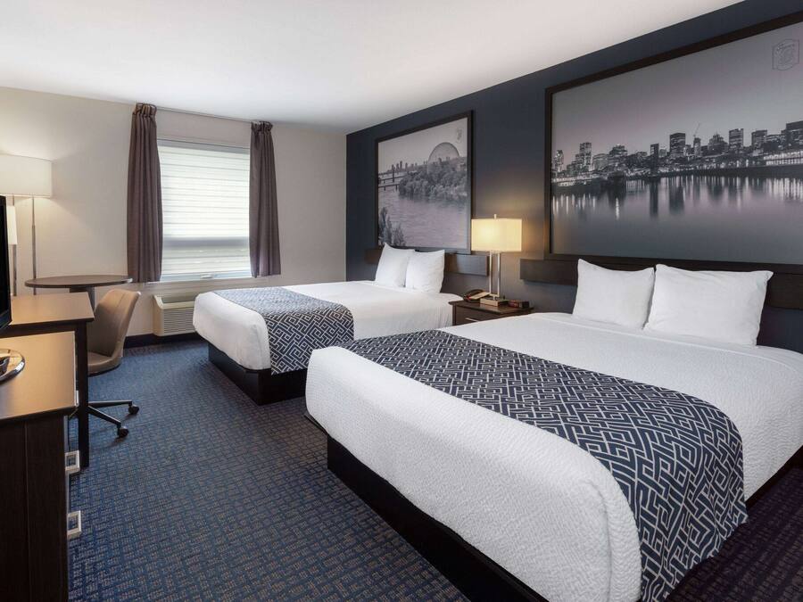 A hotel room with two beds, a desk, a chair, a TV, and a cityscape painting on the wall.