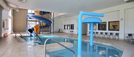An indoor swimming pool with a water slide, poolside chairs, and a clear view of the pool area.