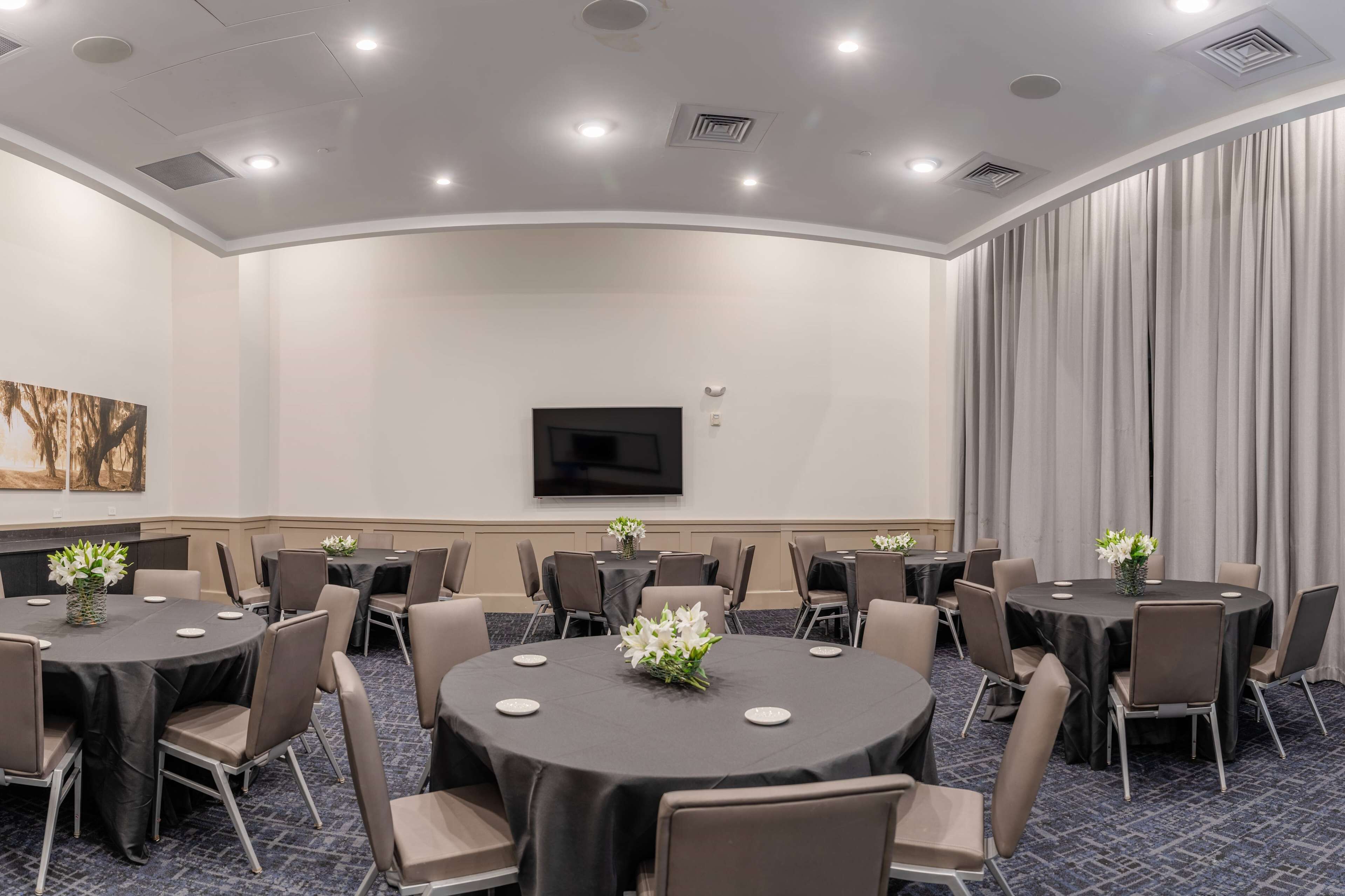 Ein Konferenzraum mit mehreren runden Tischen, jeder Tisch mit einer schwarzen Tischdecke und weißen Blumen. An der Wand ist ein Flachbildfernseher befestigt und es gibt ein großes Fenster mit Vorhängen.