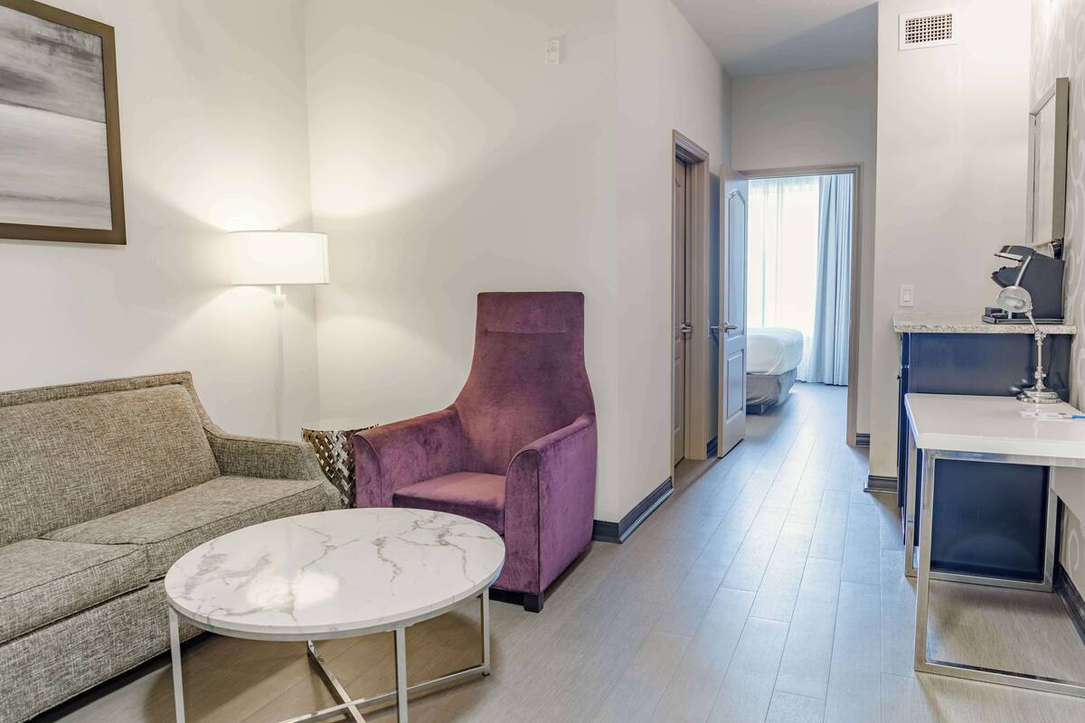 A hotel room with a sofa, a purple armchair, a round table, a lamp, a desk with a lamp, and a mirror.