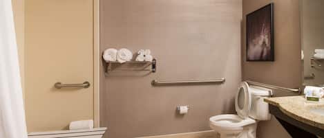 A bathroom with a shower, toilet, and sink. There are handrails, a towel rack, and a framed picture on the wall.