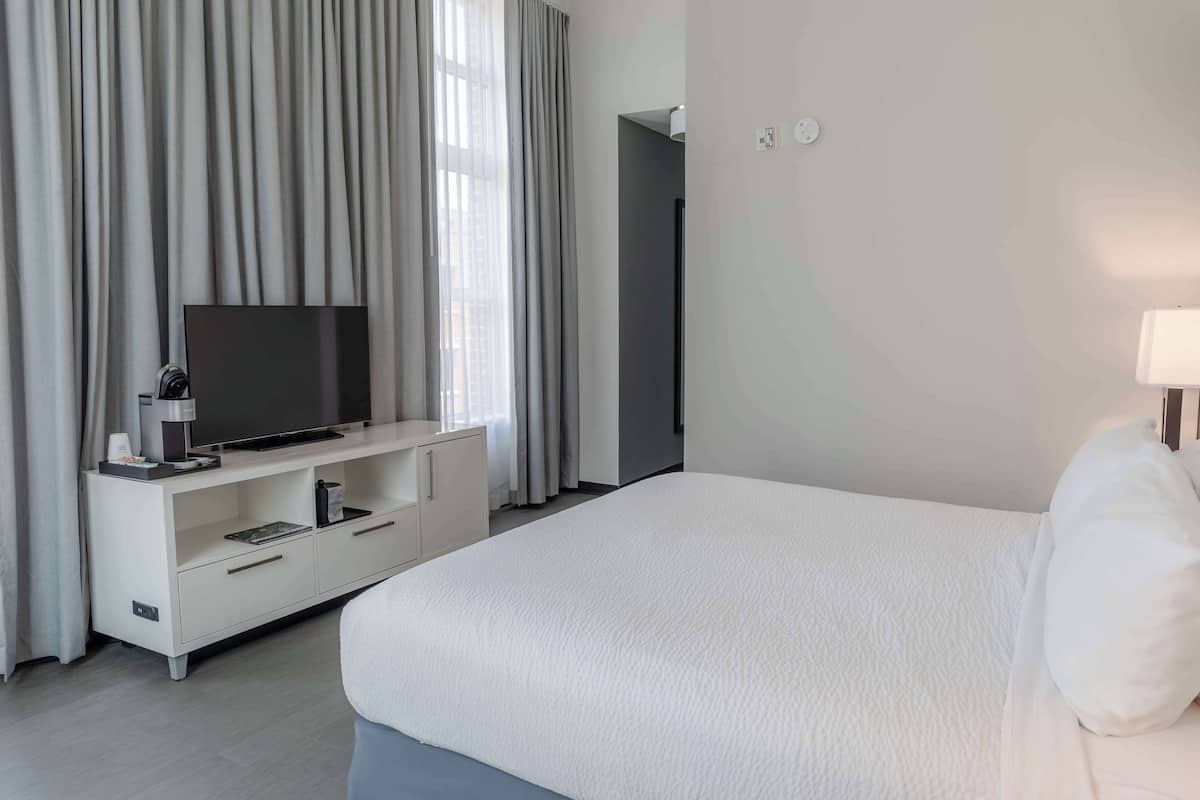 A hotel room with a large bed, a TV on a white console, and a view of the city through the window.