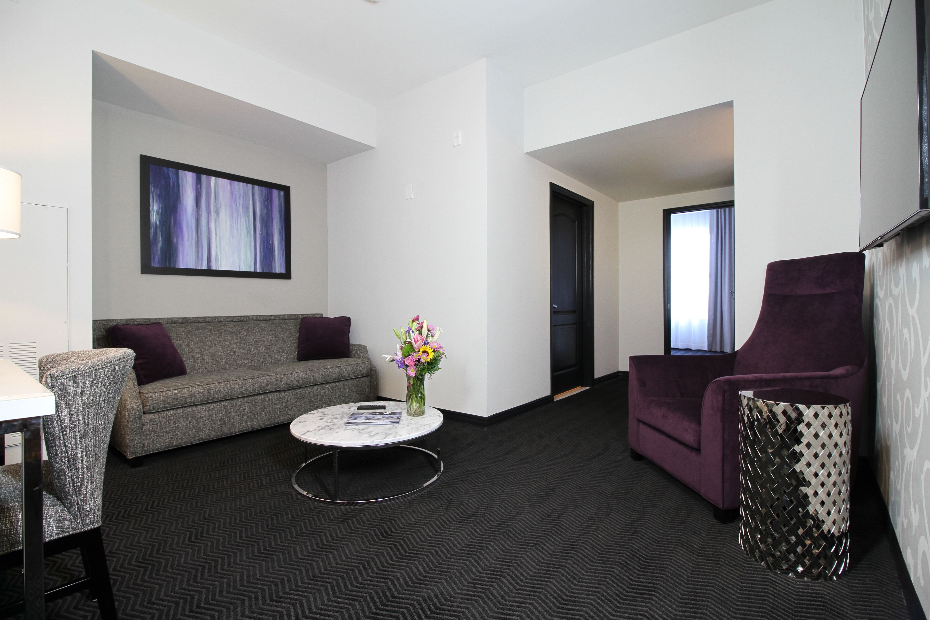 Une chambre d’hôtel moderne avec un canapé gris, un fauteuil violet, une table basse ronde et un vase décoratif avec des fleurs.