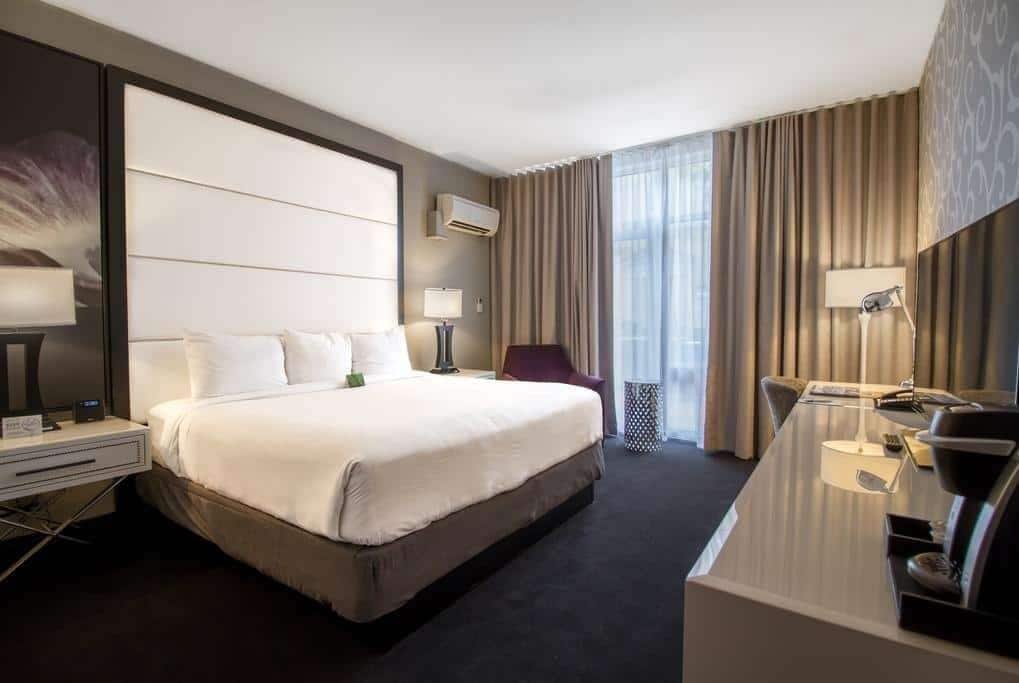 A modern hotel room with a large bed, a desk with a chair, a small table, and a lamp.