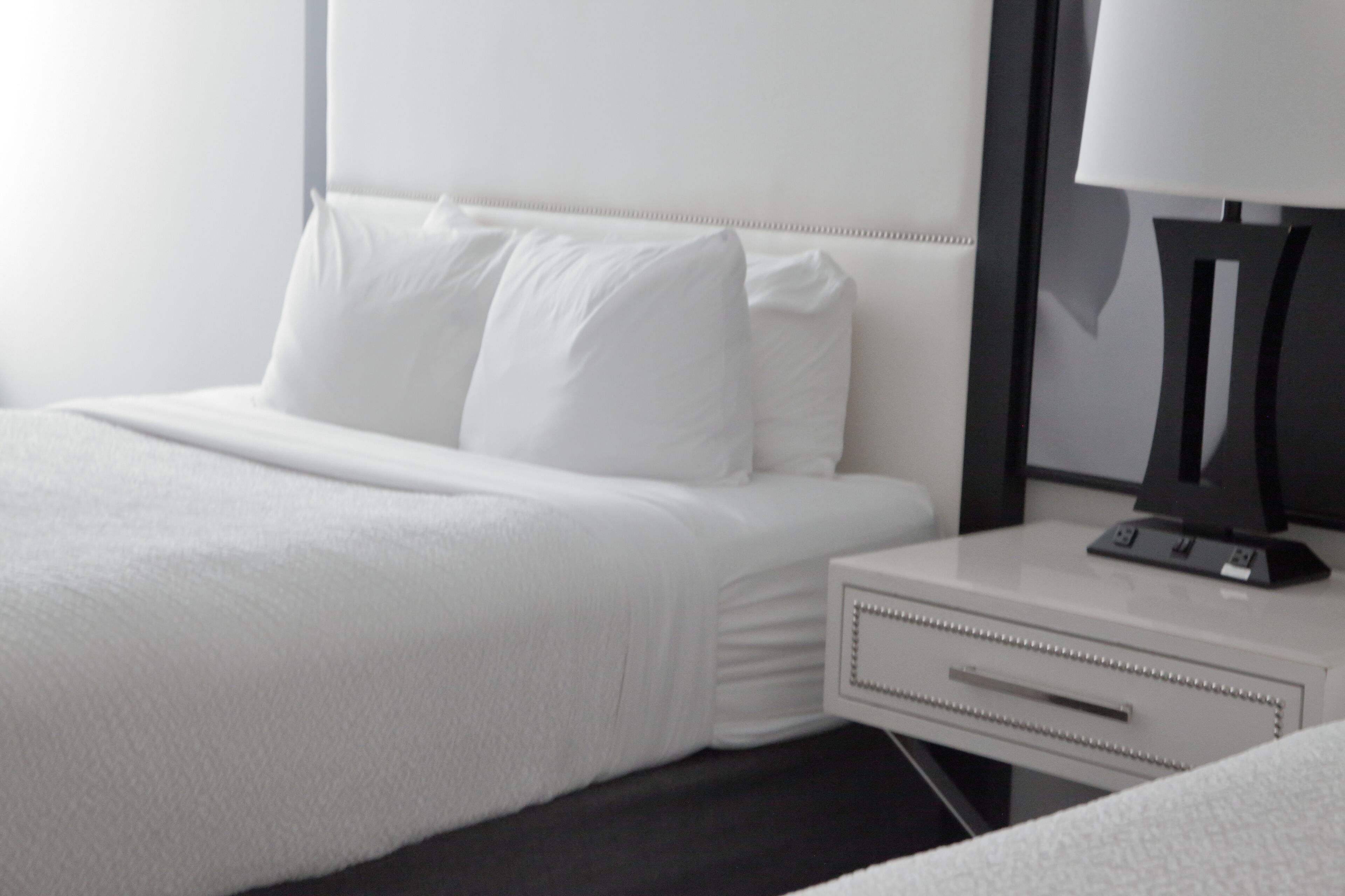 A hotel room with a neatly made bed, a bedside table with a lamp, and a drawer.