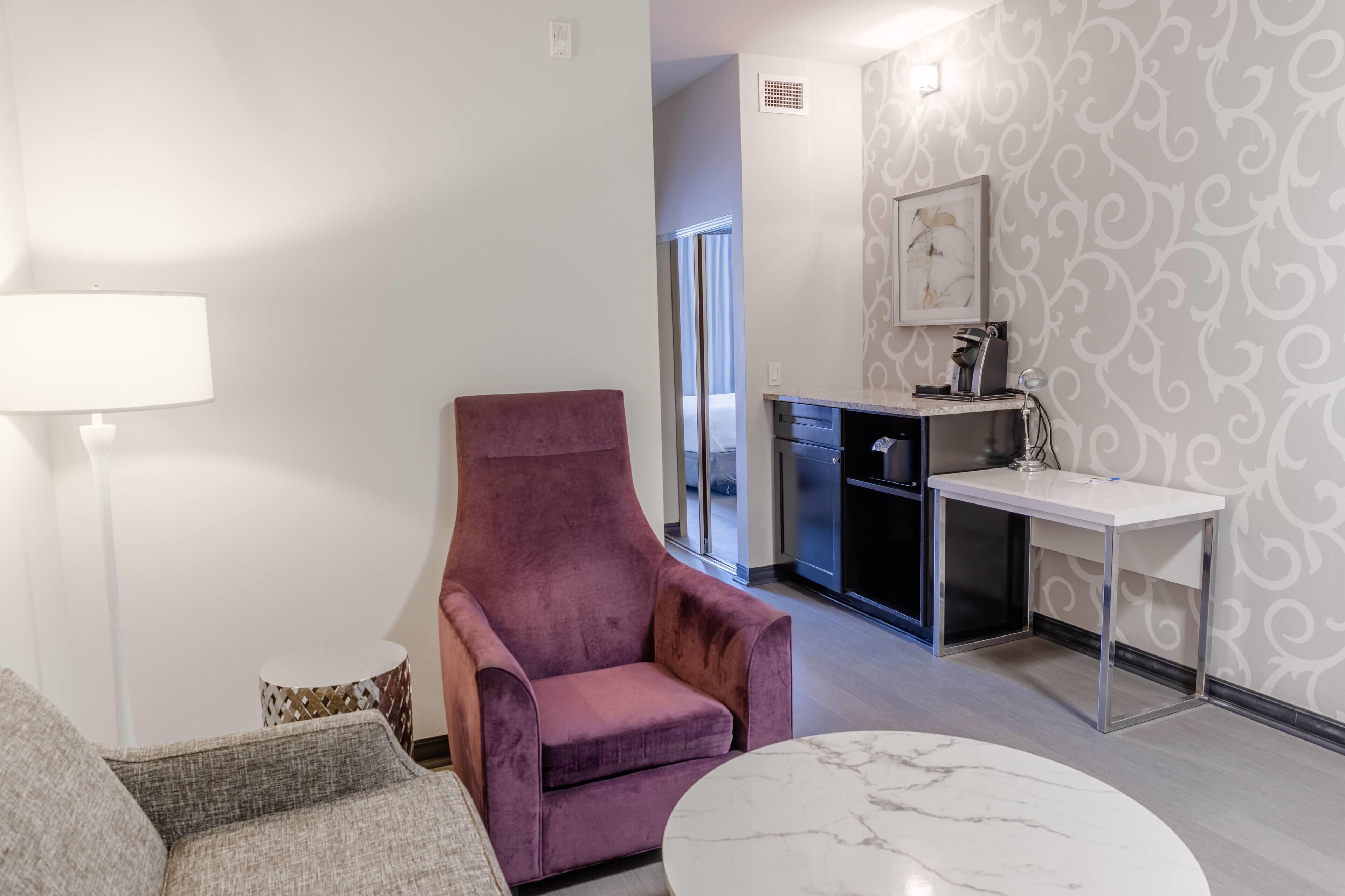 A modern living room with a purple armchair, a white lamp, a round marble table, and a kitchen area with a patterned wall and a framed picture.