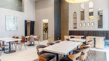 A modern dining area with high tables and cushioned chairs, a large painting on the wall, and a kitchen in the background.