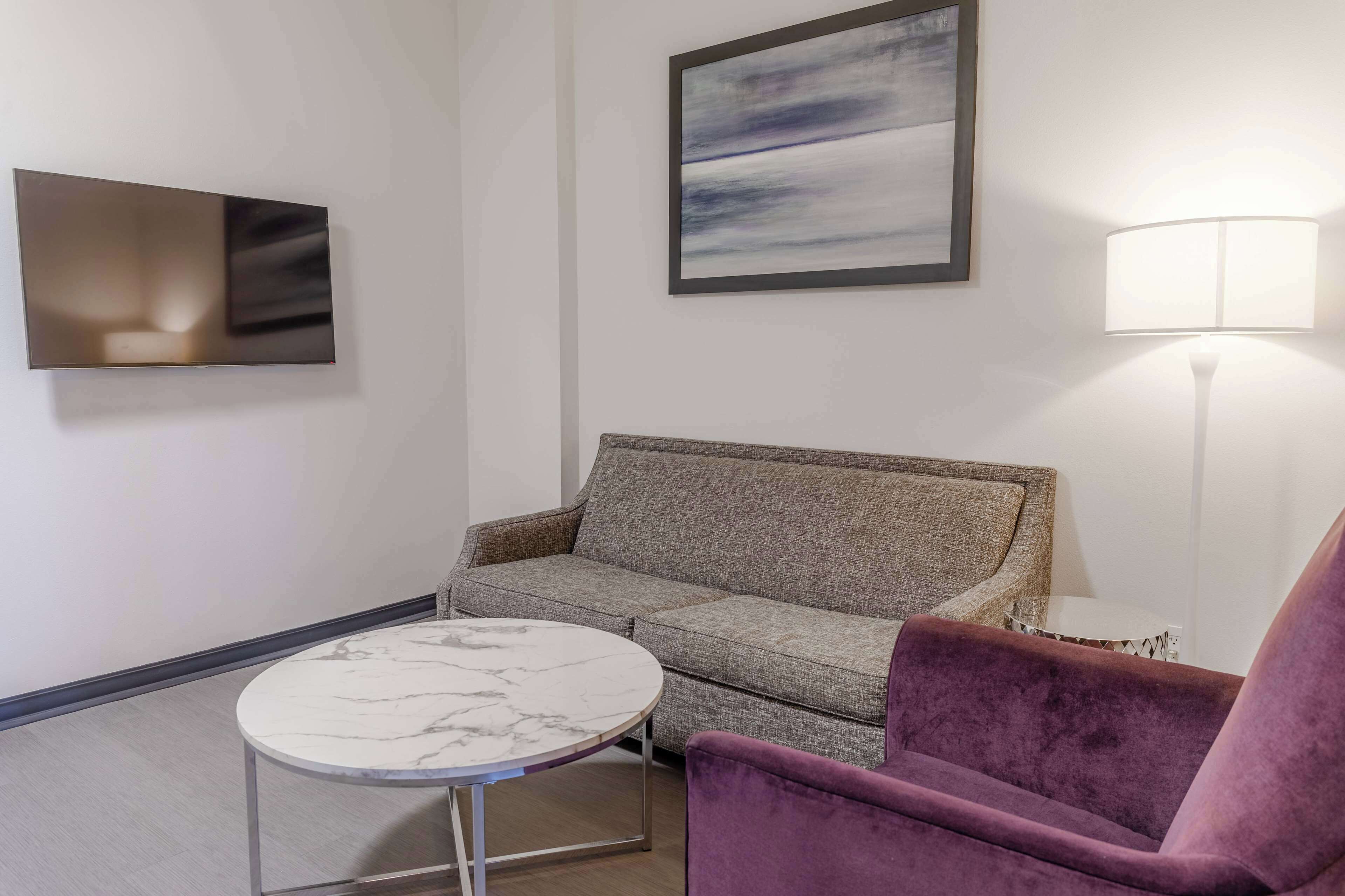 Un salon moderne avec un canapé gris, une table ronde en marbre, un fauteuil violet, un lampadaire et un tableau abstrait encadré accroché au mur.